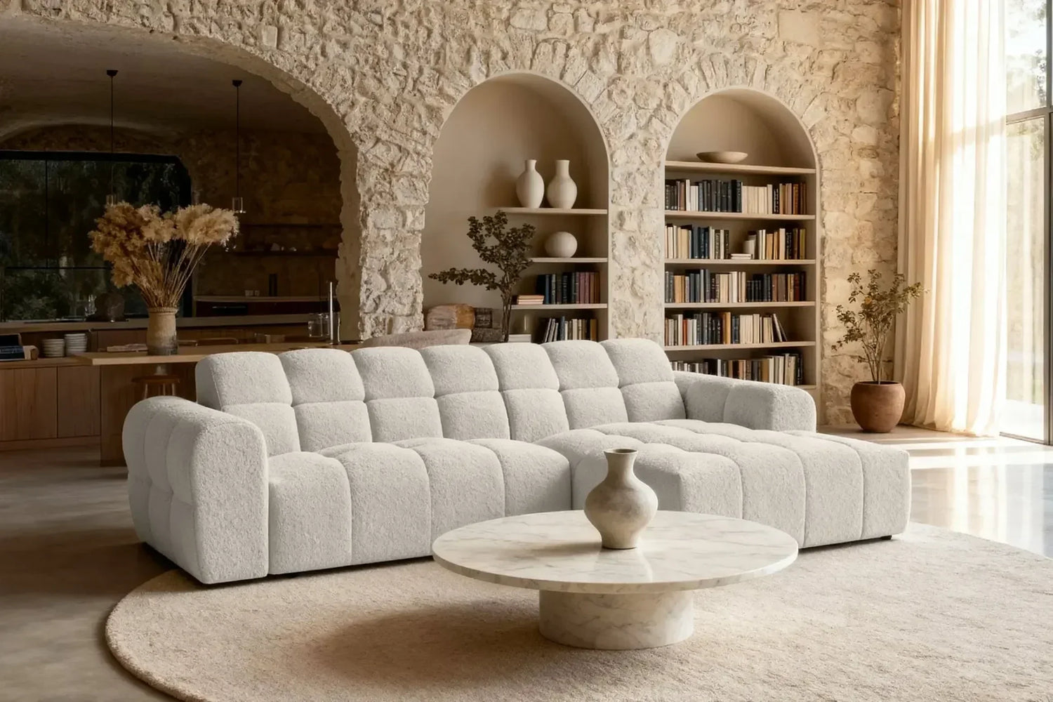 Canapé d'angle, modèle alexia dans un salon lumineu avec un mur en pierre. le canapé est en tissu bouclette posé au milieu de la pièce avec un tapis rond design